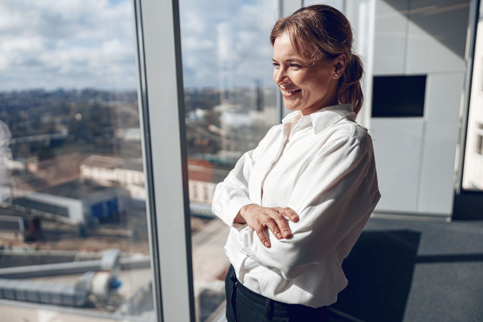 Lächelnde Frau steht mit verschränkten Armen am Bürofenster mit Blick über die Stadt