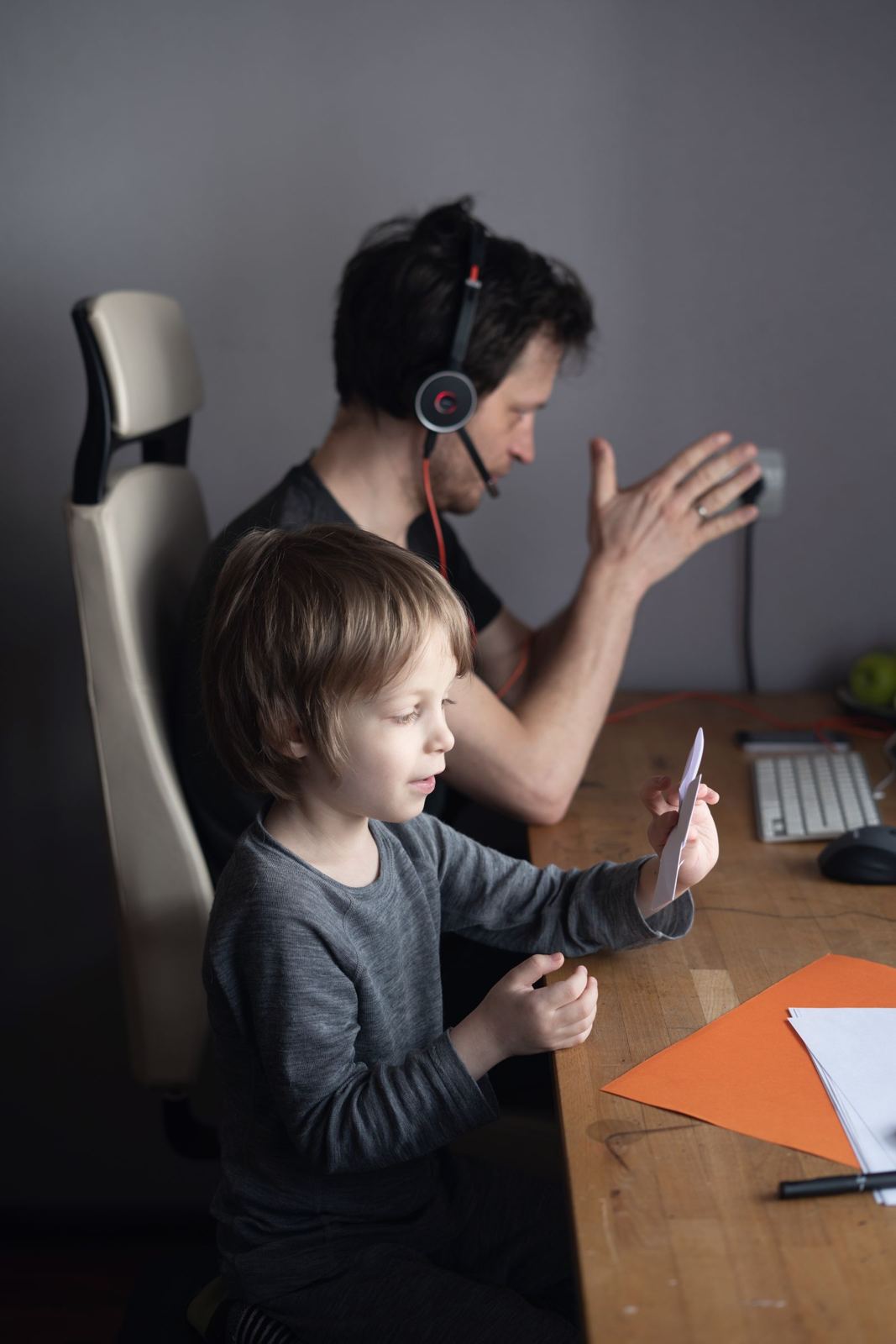 Vater arbeitet mit Headset am Schreibtisch, sein Sohn sitzt mit einem Tablet neben ihm